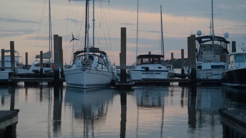 At sunrise boats sit quietly docked in a marina as soft reflections ripple across the water and a person in an orange coat passes by in the background