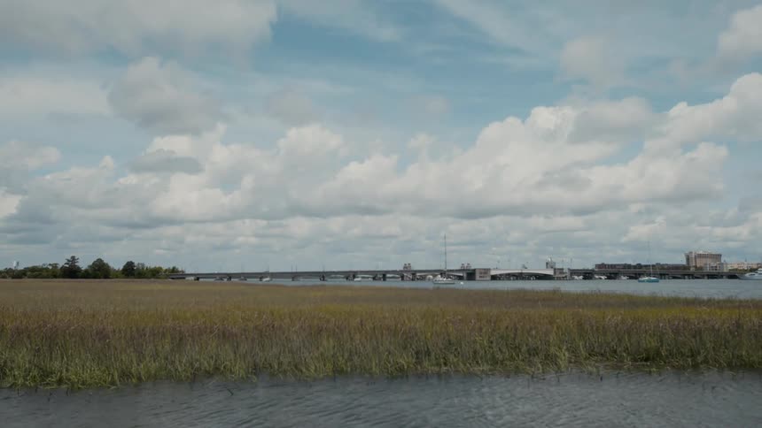 A calm marshland stretches toward the horizon where a bridge and anchored boats line a broad waterway beneath a sky filled with soft clouds
