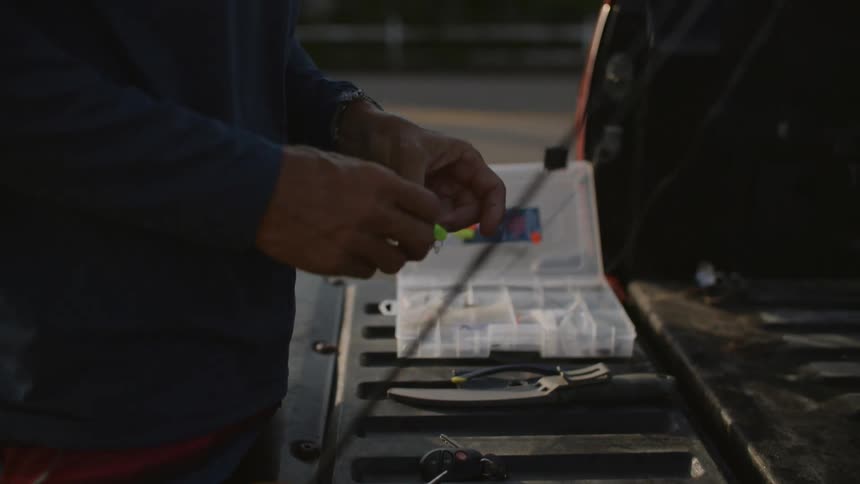 A closeup of a man at the back of a truck preparing a fishing lure and line on a fishing rod in the morning