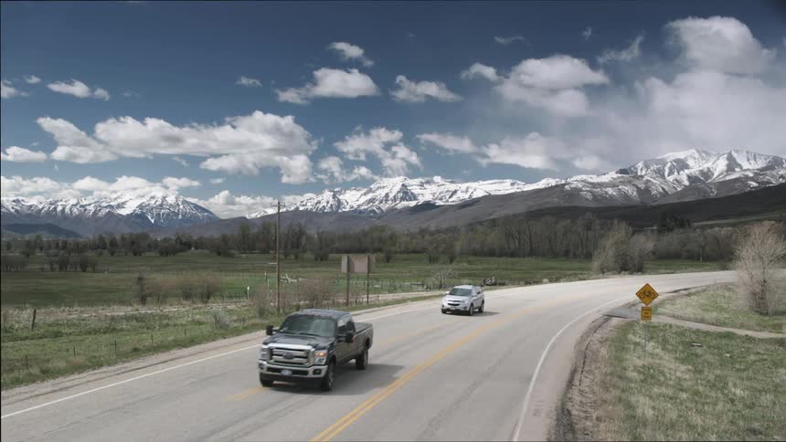 Cars drive on the countryside highway with snow capped mountains in the background