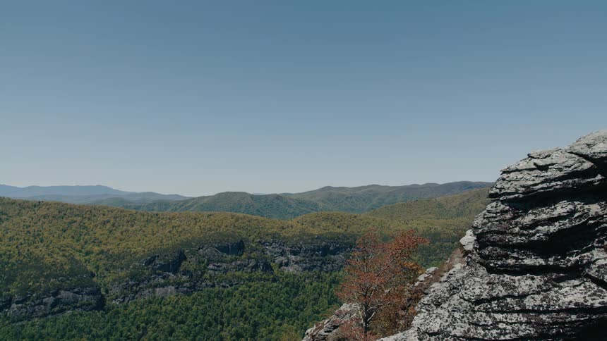 A vast mountain range covered in lush greenery extending into the distance under a clear blue sky with a rocky outcrop in the foreground