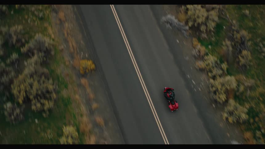 A red motorcycle rides along a deserted straight road surrounded by dry grass and scattered shrubs