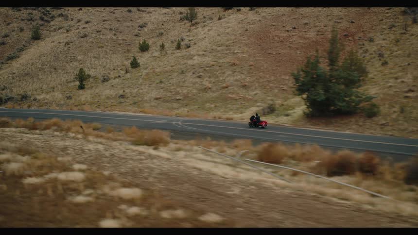 A lone motorcycle with a red storage case travels along a winding asphalt road marked with double yellow lines through dry sparsely vegetated hills under an overcast sky