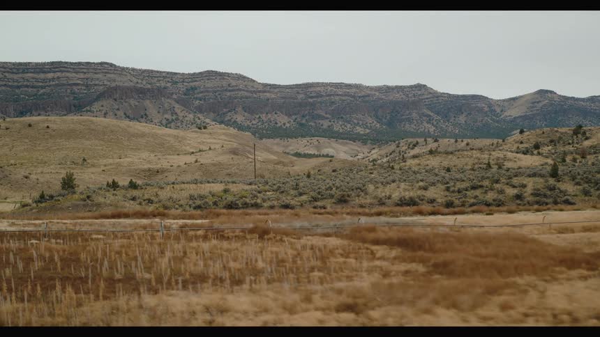 A vast stretch of golden grassland unfolds toward layered mountain ridges under a cloudy sky with a long irrigation pipe running through the field