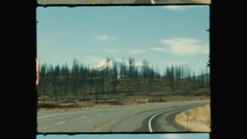 Two dark vehicles driving on a winding highway that stretches towards a majestic snow capped mountain range under a clear blue sky with scattered clouds
