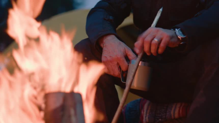 A man sitting by a bright roaring fire holding a white mug in their left hand and a thin wooden stick in their right while wearing a dark jacket blue jeans and a watch on their left wrist