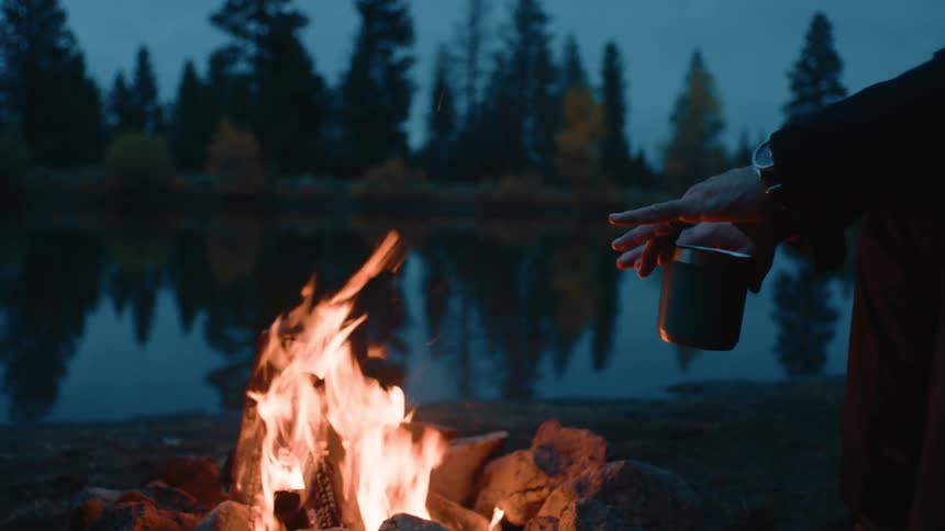 A man is warming himself by a lakeside campfire