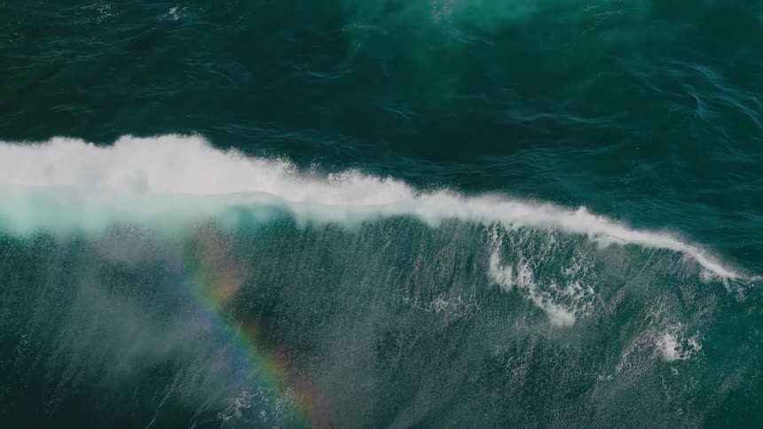 Aerial view of a large wave breaking in the ocean with white foam and a faint rainbow visible against the deep blue water