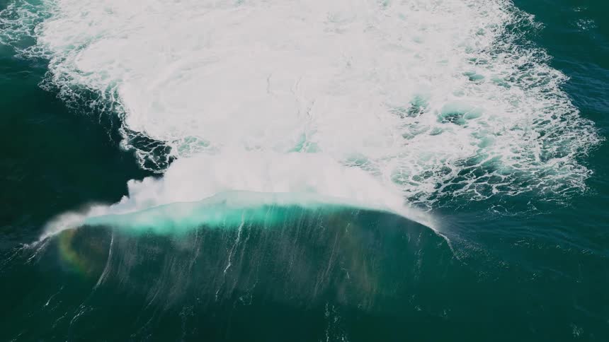 An aerial view shows a large foamy wave crashing in the deep blue ocean creating a dynamic contrast between the white foam and the dark water