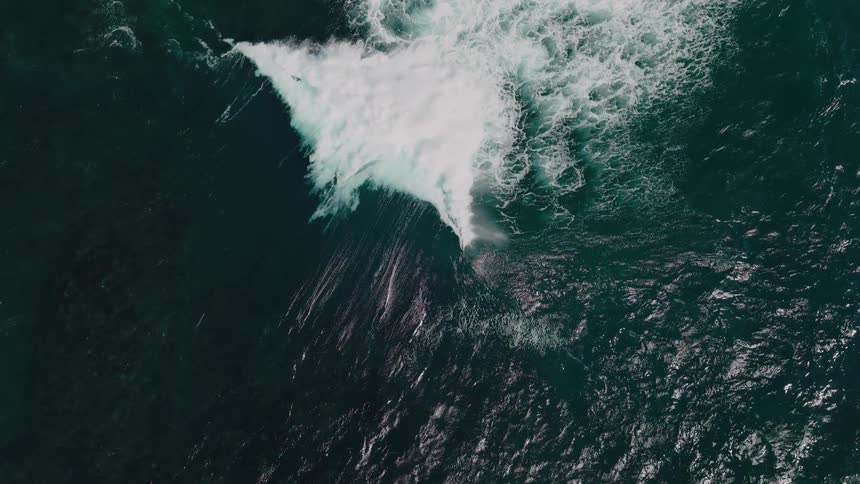 An aerial of a powerful wave crashing into the deep blue ocean creating a burst of white foam that contrasts sharply with the dark water