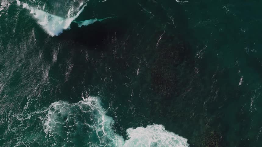 An aerial view shows a large white wave crashing in the deep blue ocean creating a dynamic contrast and texture against the dark water