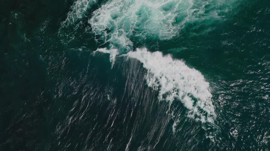 An aerial view shows a wave crashing into the ocean creating a white foamy streak against the deep blue green water