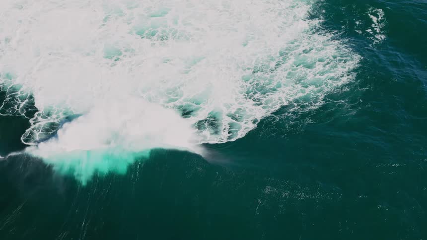 An aerial view of a large wave breaking its white foam contrasting sharply with the deep blue ocean with a hint of a rainbow visible in the spray
