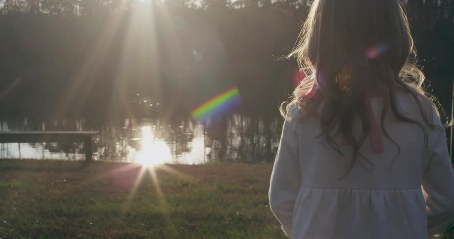 A little girl is facing a pond where the sun's rays are reflecting in the water