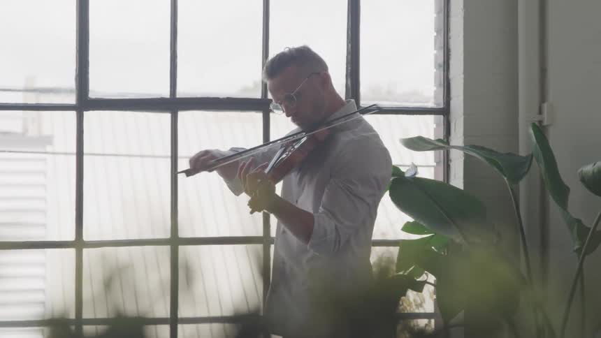 Man playing the violin next to a large window with grids in an apartement setting