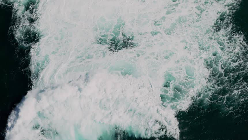 An aerial view shows a large foamy wave churning in the ocean creating a swirling pattern of white and turquoise against the dark water