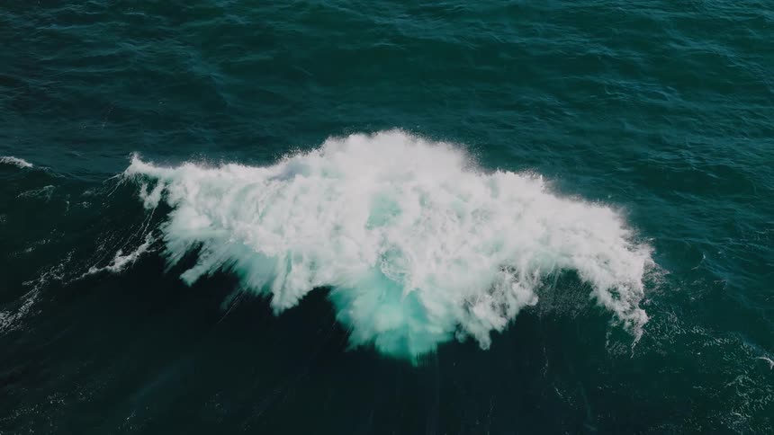 A large wave breaking in the deep blue ocean creating a burst of white foam that contrasts sharply with the dark water