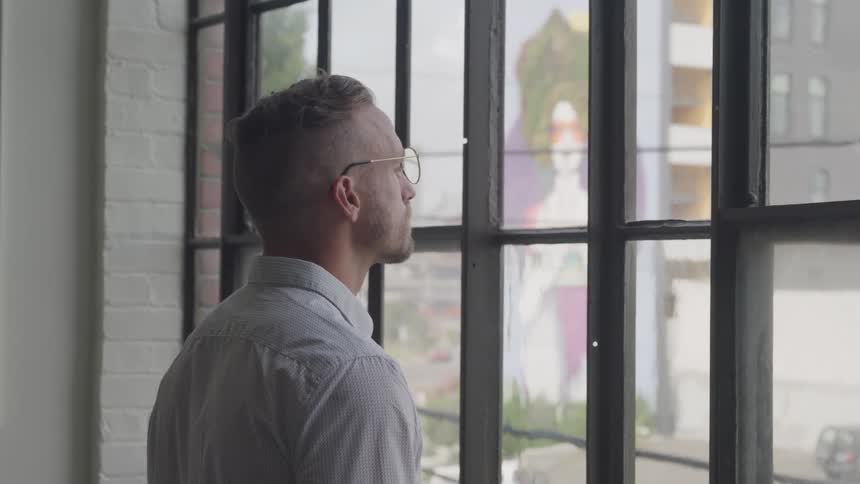 A man wearing glasses is thinking while gazing out the office windows