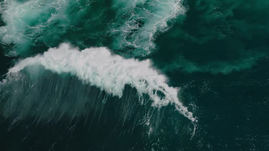 Aerial view of a large wave crashing into the ocean creating a white foamy streak against the deep blue green water