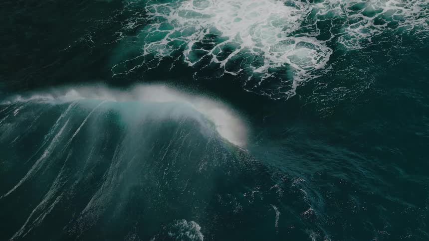 An aerial view shows a wave crashing into the ocean with white foam and a faint rainbow visible against the deep blue water