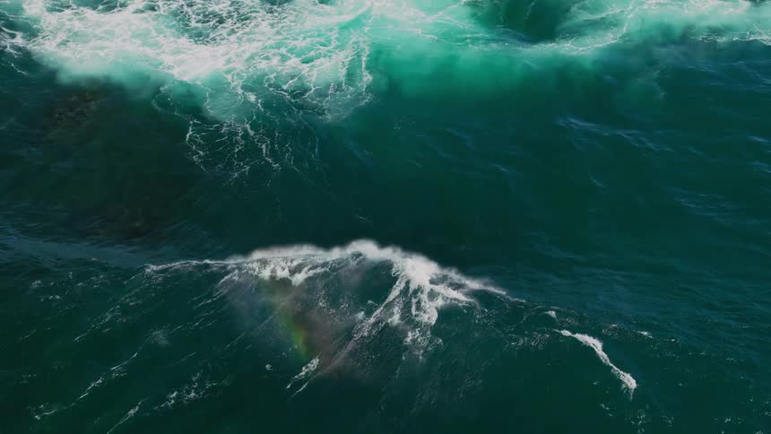 An aerial view of the dramatic interplay of dark churning ocean waves and white foam with a faint rainbow visible in the spray showcasing the raw power of the sea