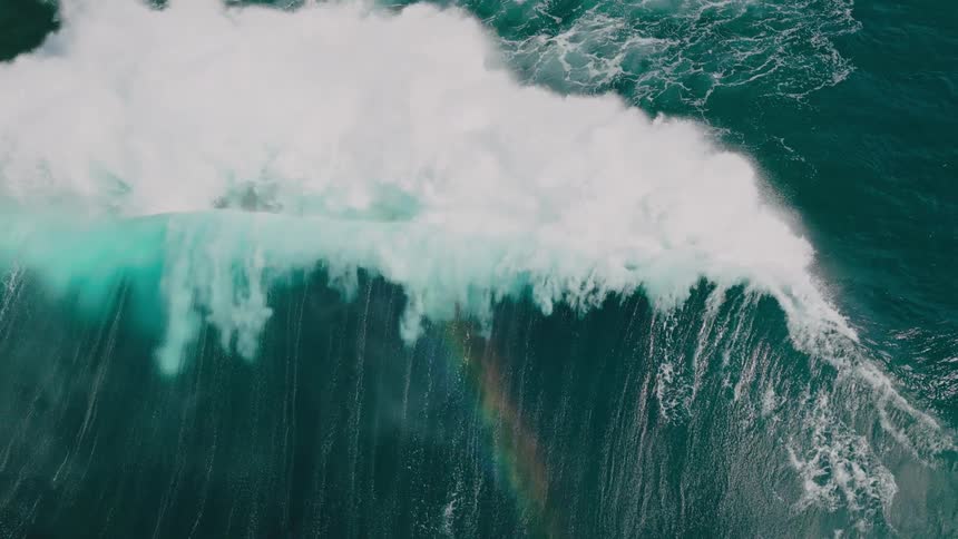 An aerial view of a large wave with foamy peaks leaving a rainbow trail in its wake
