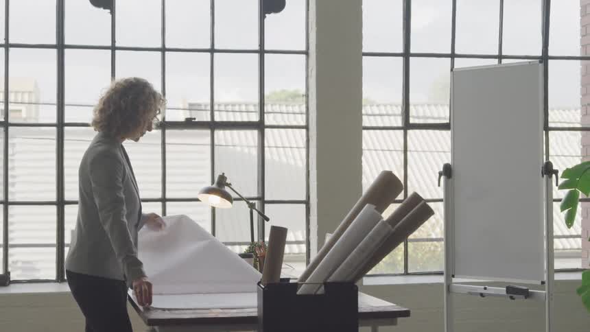 Female architect looking over blueprints in her office next to a large window with grids