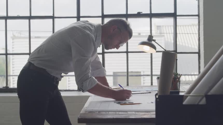 A person stands at a drafting table pen in hand meticulously working on detailed architectural plans under the glow of a desk lamp in a spacious bright room