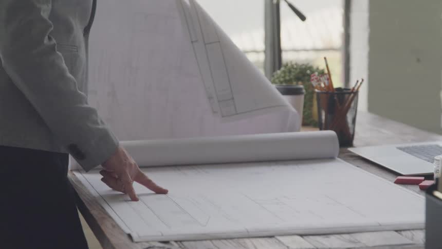 A person inspects a large architectural blueprint onto a wooden desk in a bright workspace