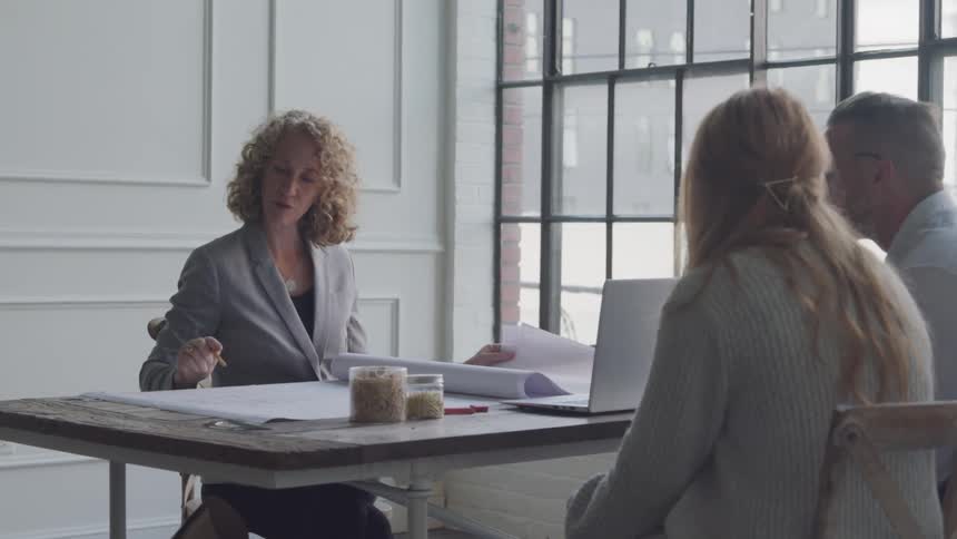 A woman is going through plans on broadsheets of paper with two people on the other side of a table who may be her clients