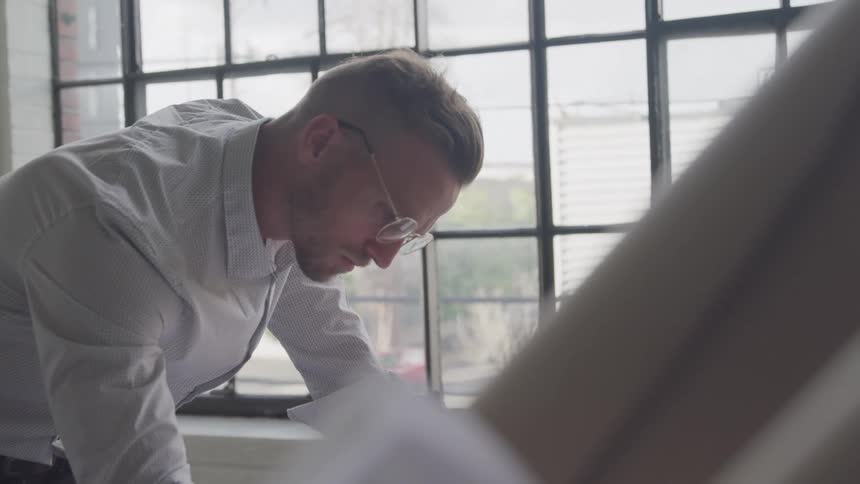 A person leans over a table near a large window seemingly focused on something placed on the table