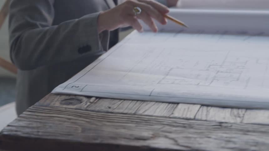 A person holding a pencil inspects architectural plans spread across a wooden surface