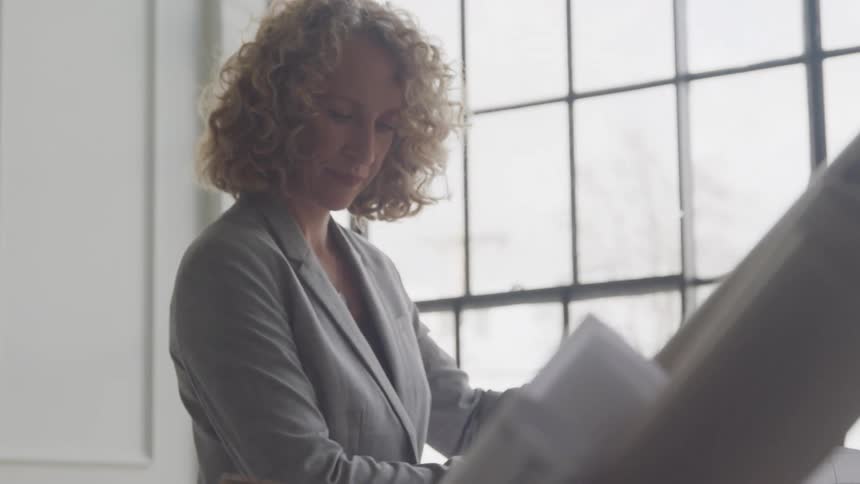 A woman in a formal blazer inspects architectural blueprints beside large windows on a bright day
