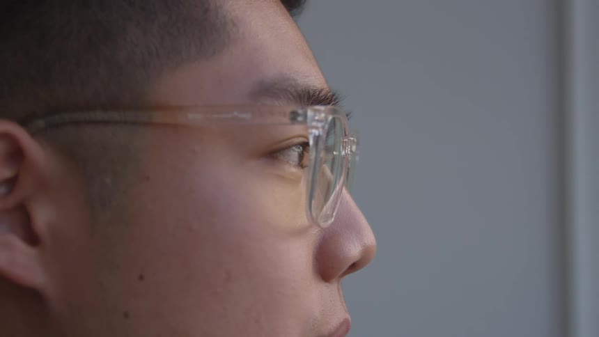 A closeup profile of a man wearing clearframed glasses gazing thoughtfully out of a window
