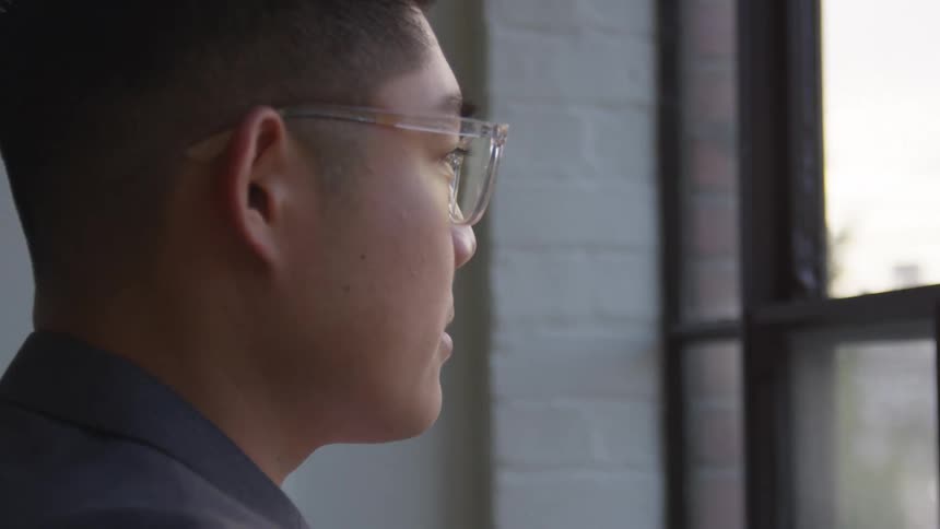A man wearing glasses and a suit looks pensively out of a window
