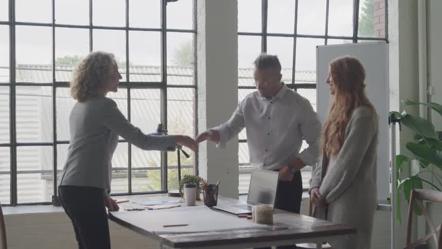 Three people are in an office setting shaking hands over a table near large windows before sitting down