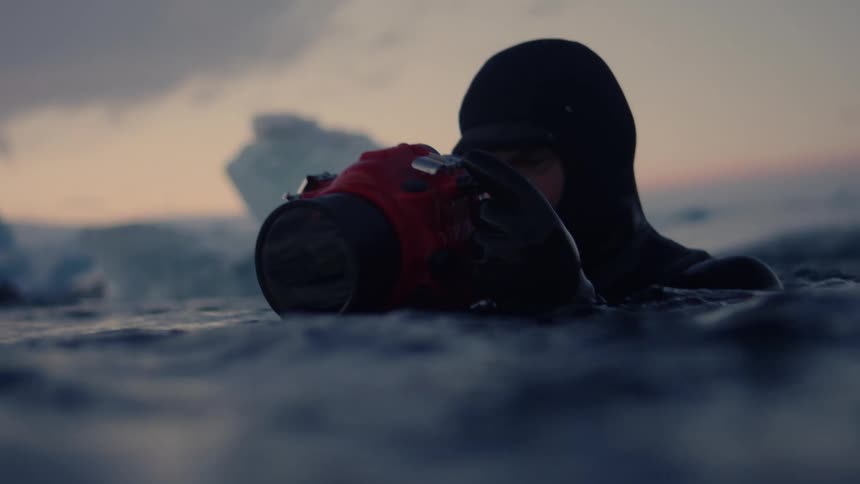 A lone diver emerges from icy waters under a dusky overcast sky with large ice formations in the background
