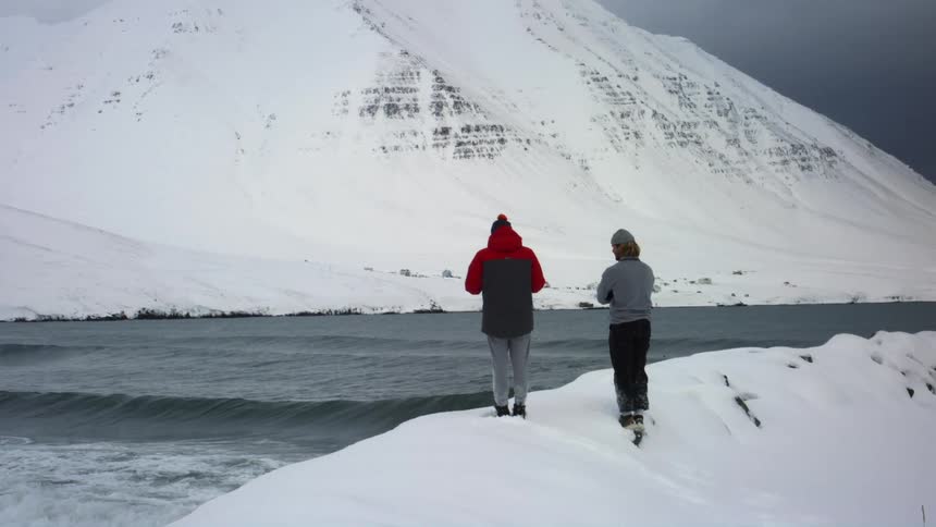 A man in a heavy winter coat and a woman in a thin grey jacket are standing on a snow bank looking out over the ocean and at a snow covered mountain across the ocean from them with the woman crossing her arms and shifting her weight to stay warm