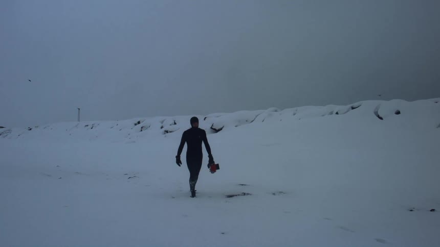 A person in a snowsuit walks through a snowy tundra while holding a camera