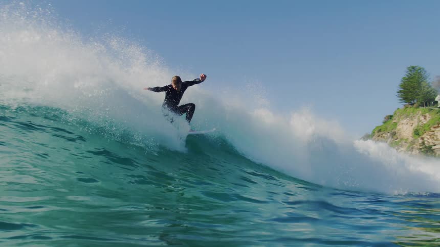 A surfer in a dark wetsuit rides a breaking wave under a clear bright sky