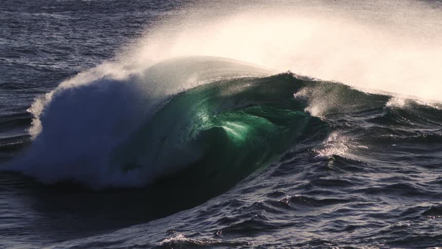 A powerful ocean wave curls and crashes creating a burst of white foam against the dark surface of the water