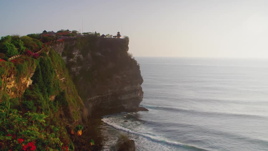 A dramatic cliffside view captures a temple perched atop a rocky promontory overlooking the ocean with waves crashing against the base of the cliffs