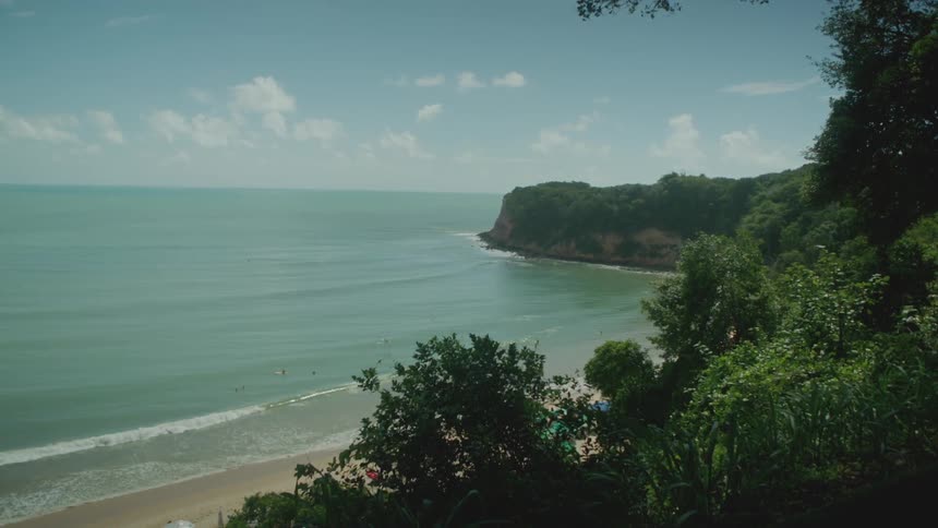 Blue calm ocean with calm waves lapping against the beach the beach is surrounded with lush green trees and shrubbery the sky is blue with scattered clouds on a bright sunny day