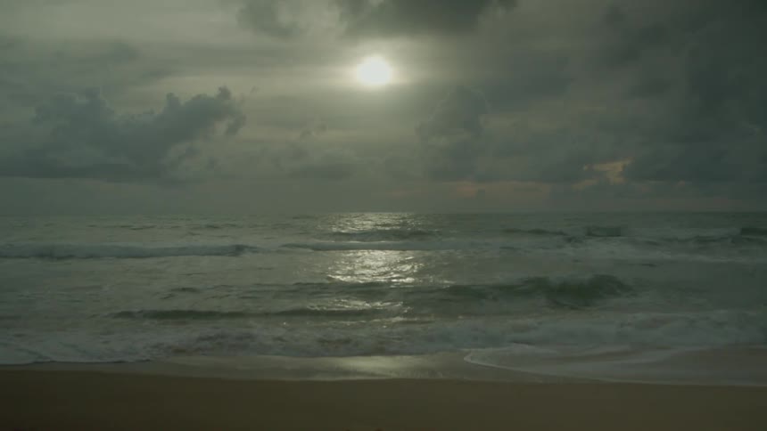 A moody seascape captures a beach with gentle waves under a cloudy sky where the sun struggles to break through creating a diffused ethereal light