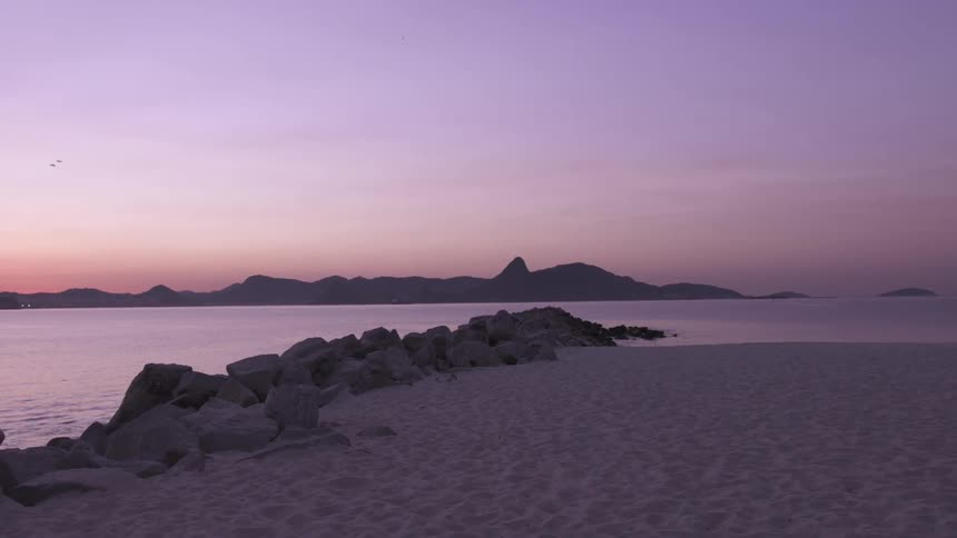 A serene twilight scene captures a rocky breakwater extending into calm waters with silhouetted hills in the distance under a pastel colored sky