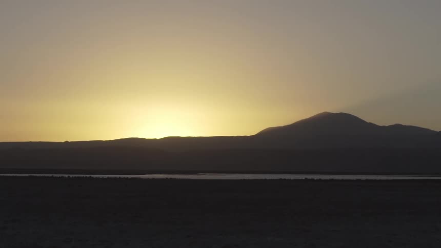 A sunset silhouette shows a mountain range against a pale sky with a body of water and dark foreground visible