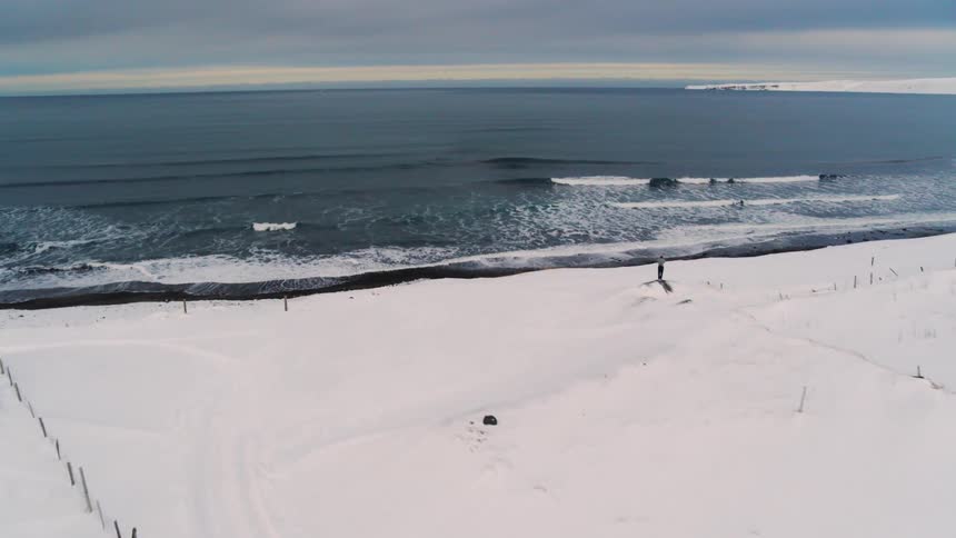 Person stands on a hill overlooking a black sandy beach in a fenced pasture on a snowy winter day