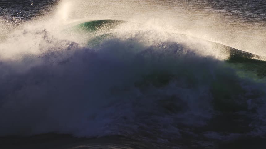 A large dark green wave curls and breaks its crest illuminated by sunlight creating a dynamic contrast between the shadowed water and the bright foam