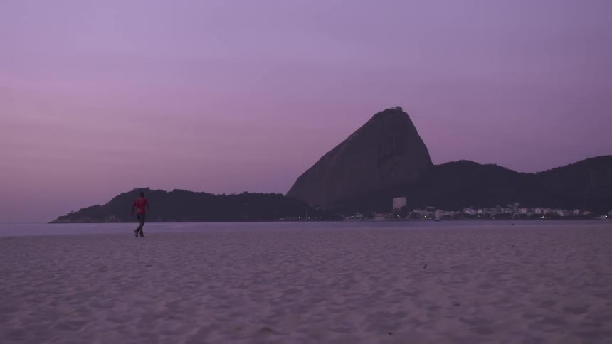 Person jogs slowly across the beach while a coastal town nestles near mountains in the background