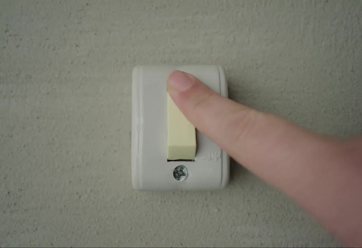A finger presses a light yellow toggle switch mounted on a textured lightcolored wall